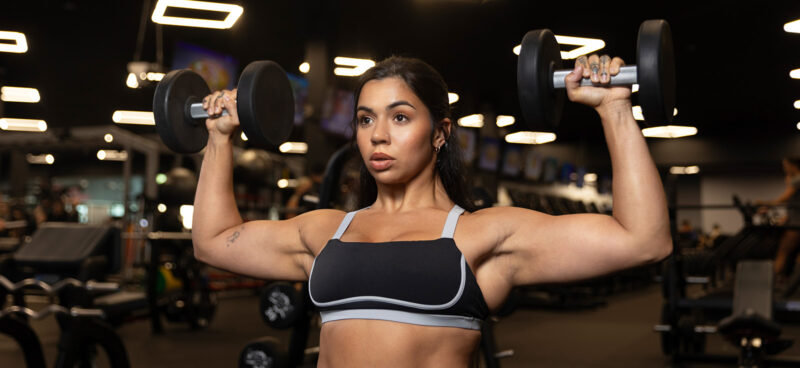 A woman is doing a seated dumbbell shoulder press in a gym, holding a dumbbell in each hand at shoulder height with her elbows bent and arms out to the sides. She’s wearing a black and gray sports bra, and the shot is framed closely on her upper body, highlighting her shoulders and arms. The background is softly blurred with gym lights and equipment visible.