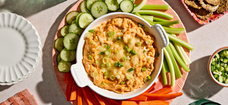 Overhead view of a creamy baked buffalo-style dip in a white oval baking dish, topped with melted cheese and sliced green onions. The dish sits on a pink platter surrounded by sliced cucumbers, celery sticks, and red bell pepper strips for dipping. Bright sunlight casts shadows across the table, giving the spread a fresh, colorful appetizer look.